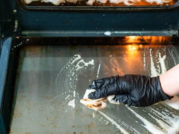 cleaning oven in home kitchen - female hand cleans glass door of dirty oven with reagent