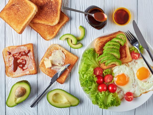 healthy breakfast - fried eggs, fresh salad, toasts with yeast spread and sliced avocado on a white plate on a wooden table, view from above