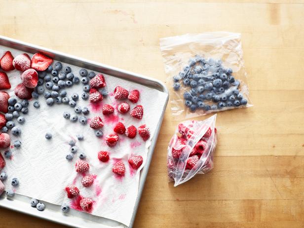 Overhead of freezing berry preparation, rasperries, blueberries and strawberries sit frozen on a paper towel on a silver baking sheet with plastic bags holding more frozen berries sitting next to the tray on a wooden surface.