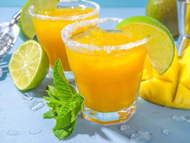 Cold summer cocktail, mango margaritas with tequila, salt lime slices, crushed ice and mint. Seasonal refreshing drink, on bright blue sun lighted background