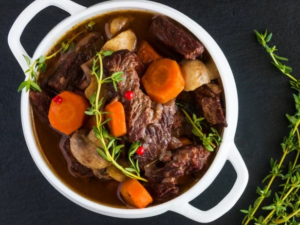 Beef Bourguignon in a white soup bowl on black stone background, top view. Stew with carrots, onions, mushrooms, bacon, garlic and bouquet garni. The dish is served with fresh thyme.