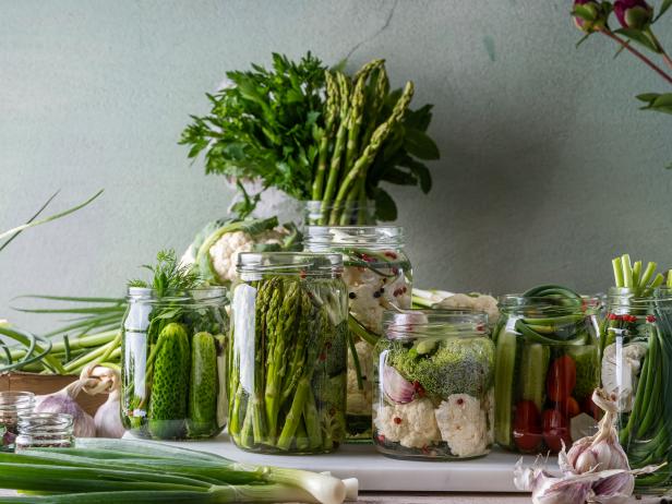 Canning process. Fresh vegetables in glass jars on rustic wood table. Garlic scapes, asparagus, cauliflower, broccoli, tomatoes, cucumber, garlic, herbs, seasoning. White and green, fermented foods
