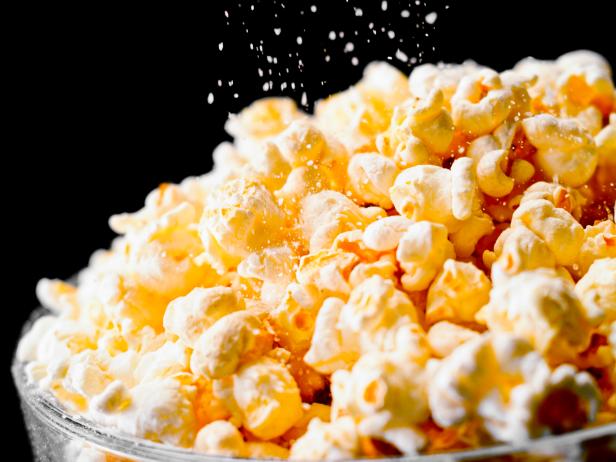 Close- up of popcorn in a glass bowl being sprinkled with salt against black background.