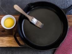 Cast iron skillet being brushed with olive oil to prevent sticking, on a slate countertop with a red dish towel