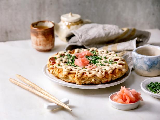 Homemade japanese fast food okonomiyaki cabbage pancake decorated by spring onion, pickled ginger, mayo sauce on ceramic plate with chopsticks and ingredients above. White marble table. Flat lay