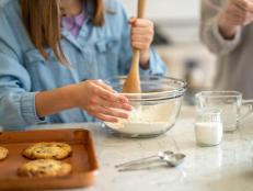 A photo showing a young girl's hands using a wooden spatula to mix her cookie batter. She is at her kitchen at home and is baking chocolate chip cookies.
