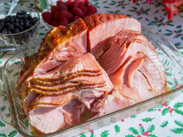 This photo shows a Christmas ham that is sliced and put on a serving dish. The cooked ham is in a pyrex serving dish and has bowls of berries and a plate of cheese in the background. The table is brightly lit and covered with a festive Christmas tablecloth with a holly and berry design. A nice and healthy eating food shot with holiday motives. - A great shot for preparation cookbooks, ingredient, catalogs, chef, diet and cooking websites or magazines.