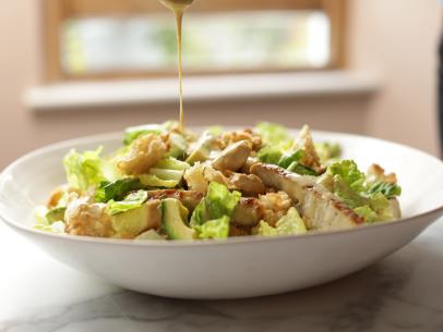Landscape, side on close-up of Caesar salad with dressing being poured overtop, as seen on Mary McCartney Serves It Up, season 3.