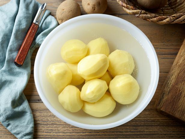 fresh raw peeled potatoes in bowl of cold water on wooden kitchen table, top view