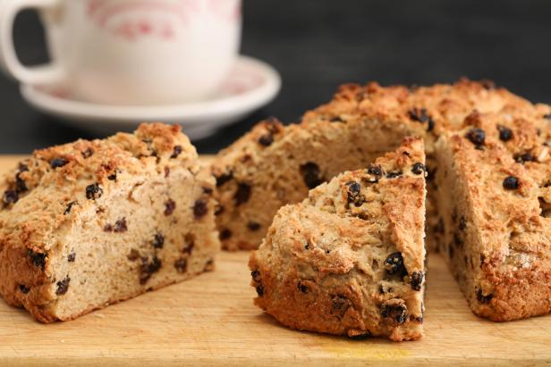 A close up horizontal photograph of a homemade,freshly baked and partially sliced loaf of Traditional, Saint Patricks Day Irish soda bread and a cup of spiked coffee or tea in the background.