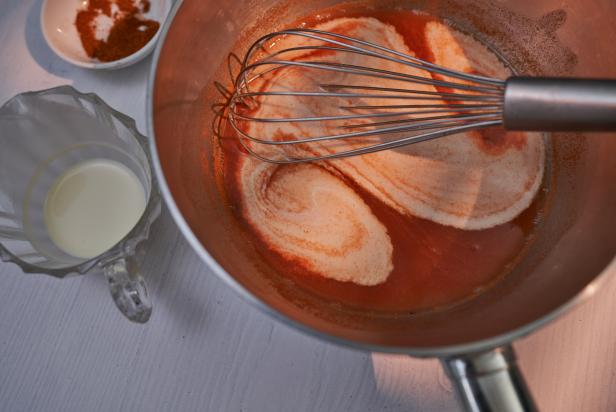 Tomato sauce preparation step. Added cayenne powder and double cream to reduced juice of sieved blended tomato chops. Whisk. Pitcher, bowl and saucepan on white wood planks. Light effect. High point of view.