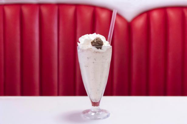 Chocolate cherry milkshake served in classic milkshake glass, topped with whipped cream, chocolate truffle and plastic straw. Positioned in retro 50s-style diner decor.