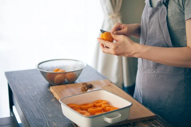 A Japanese woman using a knife in the kitchen to cut apricots in half and arrange them on an enamel bat.