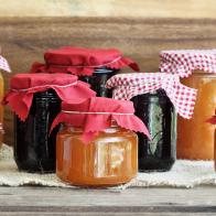 Variety of homemade jams and preserves covered with checkered and red cloth against a rustic background. Extreme shallow depth of field with selective focus on jar in front. Assortment includes peach butter, cantaloupe, blueberry, boysenberry, grape and blackberry.