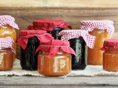 Variety of homemade jams and preserves covered with checkered and red cloth against a rustic background. Extreme shallow depth of field with selective focus on jar in front. Assortment includes peach butter, cantaloupe, blueberry, boysenberry, grape and blackberry.