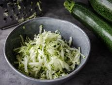 High view of a gray bowl with grated zucchini, with a grater and whole zucchinis