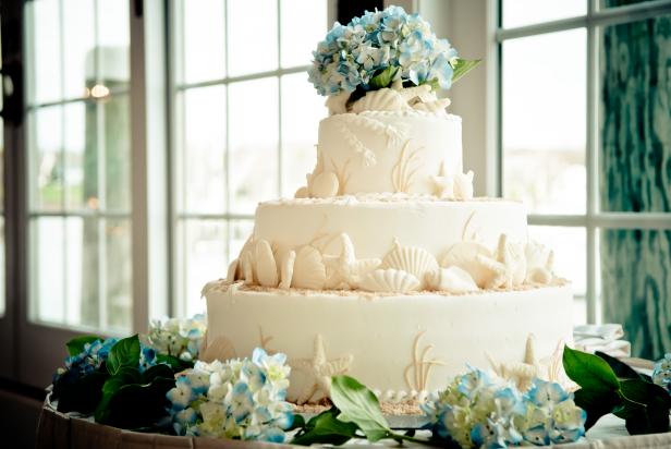 Circular wedding cake that is ocean themed. It is covered in edible shells with an appearance of sand and decorated with flowers.