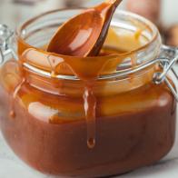Glass jar of brown salted caramel with smudges and a wooden spoon inside on a white background