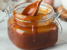 Glass jar of brown salted caramel with smudges and a wooden spoon inside on a white background