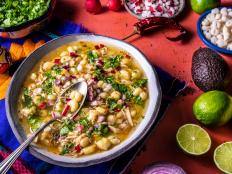 Pozole Mexican corn stew with meat, mote corn, radishes, coriander, lime and lettuce on coral pink table