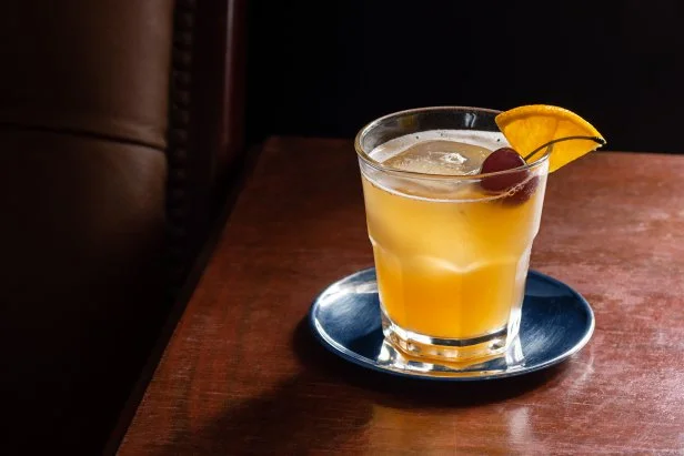 An Amaretto Sour cocktail on the rocks with a cherry and orange slice garnish on a dark wood table next to a leather seat. This drink is made with amaretto liqueur, lemon, and simple syrup (or sour mix) and topped with a cherry and orange. Taken in a dark luxurious bar or restaurant.