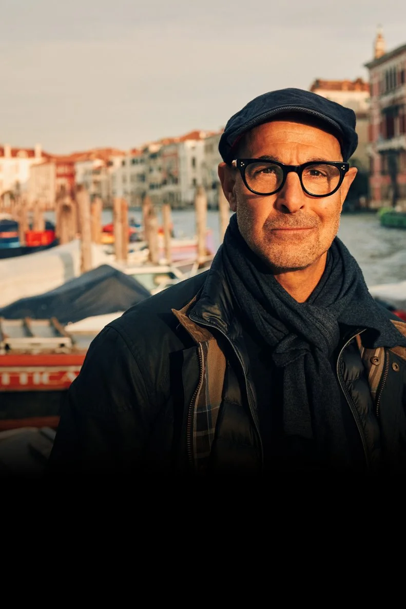Stanley Tucci posing outdoors with a scene of Italy behind him