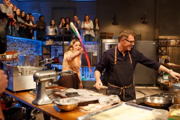 Co-host Damaris Phillips waves a ribbon at chef Bobby Flay while he tries to prepare his Round 2 dish, as seen on Beat Bobby Flay, Season 24.