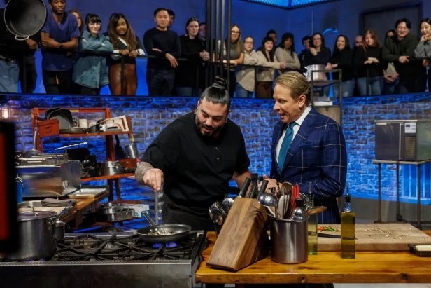 Chef Logan Sandoval sprinkles an ingredient into a pan over a lit stove as Carson Kressley speaks to him.