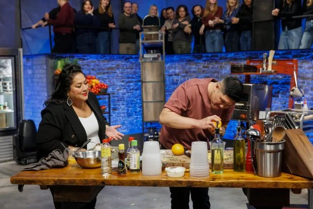 Ayesha Nurdjaja stands next to chef Laurence Louie at his cooking station watching him zest a lemon.