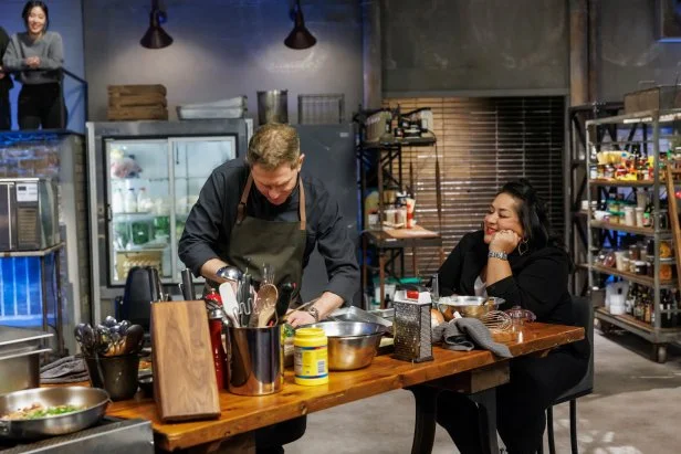 Bobby Flay keeps his head down while cooking as Ayesha Nurdjaja sits at his cooking station and watches. 
