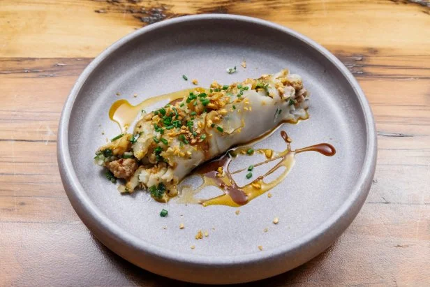 An overflowing roll wrapped in a rice noodle with ground pork and shrimp spilling out of the sides, topped with sweet soy sauce, chili oil, crunchy garlic and chives.
