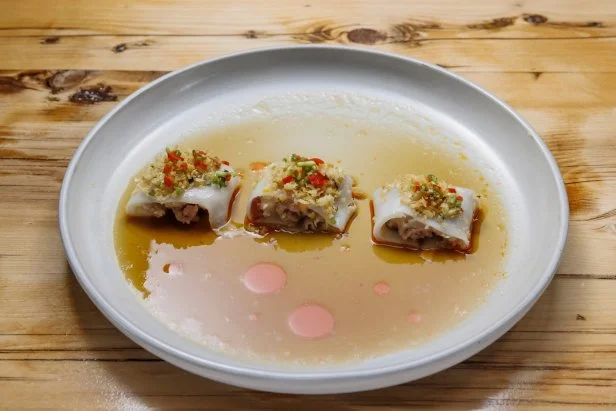 Chef Laurence Louie's Cheung Fun: a rice roll sliced into three pieces with a filling of ground pork, Chinese sausage, and shrimp topped with toasted ginger scallion panko and chilies, placed in a small pool of soy sauce.