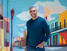 A man in a blue long sleeved shirt smiles while standing in front of a mural. 