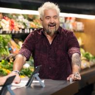 Host Guy Fieri welcomes the judges, as seen on Guy's Grocery Games, Season 39, in front of a produce display.
