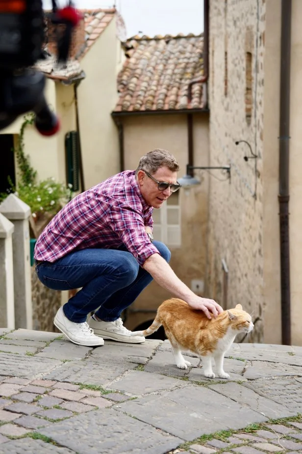 Bobby Flay pets a cat while walking through Montalcino in Tuscany, Italy, as seen on Bobby & Giada in Italy, Season 1.