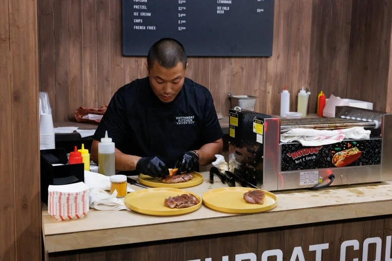 Chef Phuoc Vo cooks using a hot dog roller and a popcorn machine after being exiled to the snack bar kitchen during round two, as seen on Cutthroat Kitchen: Knives Out, Season 1.