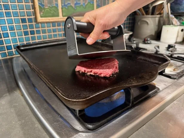 Stainless steel burger press smashing a hamburger patty on a stovetop griddle
