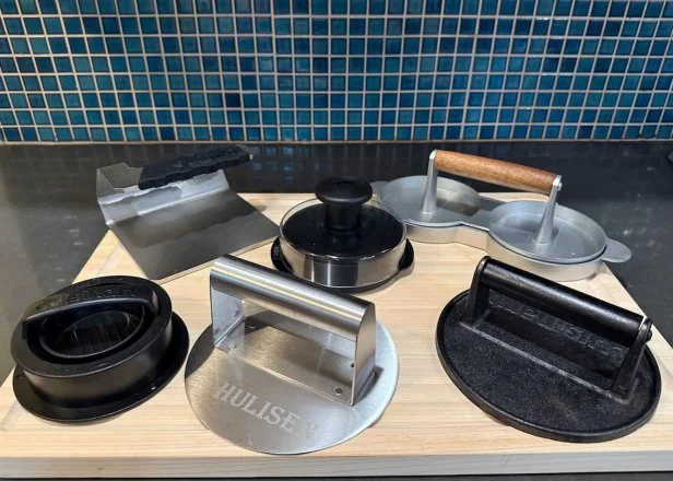 Six burger presses and smashers arranged atop a wooden cutting board