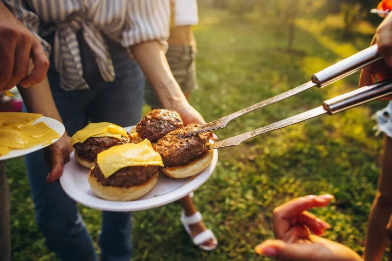 Cheese. Image of tasty homemade burger with melting cheese, tomatoes and grilled meat. Group of friends having nice outdoor picnic, making food, drinking beer and having fun.