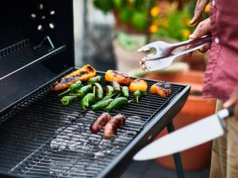 Man holding BBQ tongues turning green chillies, char grilled yellow peppers and sausages. Cooking, skill, taste, anticipation