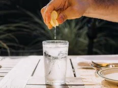A hand is squeezing a lemon slice over a glass of sparkling water with ice, set on a wooden table outdoors. Sunlight illuminates the scene, creating a refreshing and summery atmosphere.