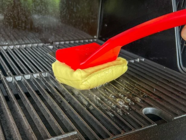 Steam grill brush with a red body and yellow sponge head cleaning a grill grate with water and steam
