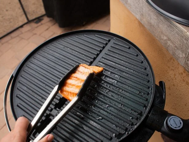 A pair of tongs flipping a piece of salmon that's cooking on a black outdoor electric grill