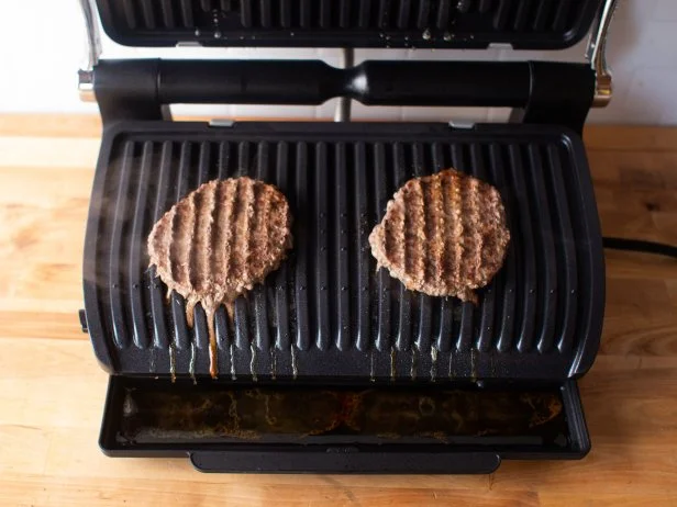 Two burger patties with grill marks cooking on a black indoor electric grill atop a wooden countertop