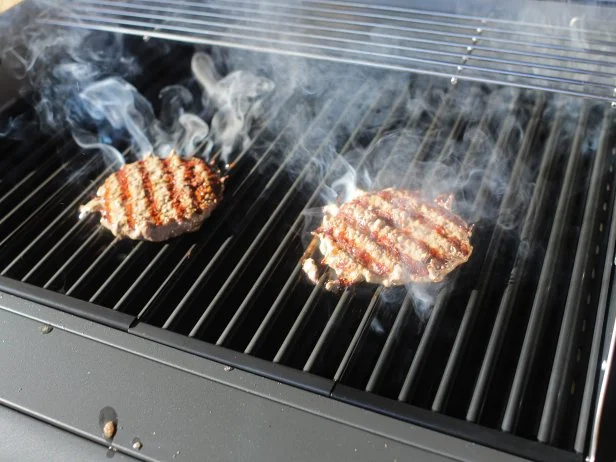 Two burger patties with grill marks cooking on an outdoor electric grill with smoke rising from the grates