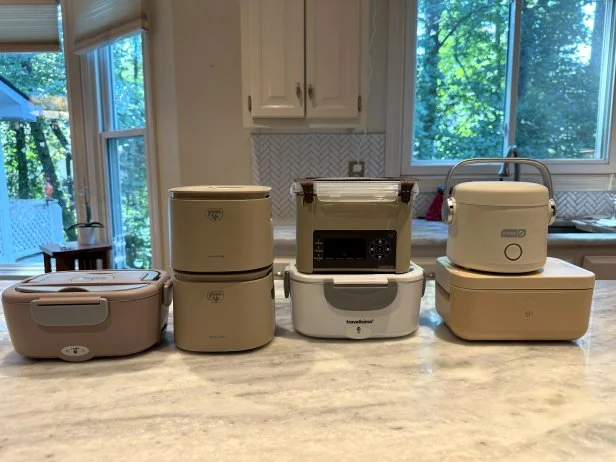 Seven electric lunchboxes in neautral colors arranged together on a white marble countertop in a white kitchen with glass windows