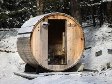 round wooden sauna outdoors covered in snow during winter blizzard snowstorm (snowy weather outside with wood barrel shaped glass door steam room) home back yard patio spa health cold relaxation