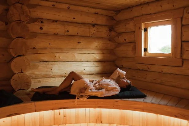 A tranquil scene in a rustic log cabin features a woman resting on a cushioned bench, illuminated by soft warm light from a small window. Calm, cozy, and restful mood for lifestyle or wellness themes.