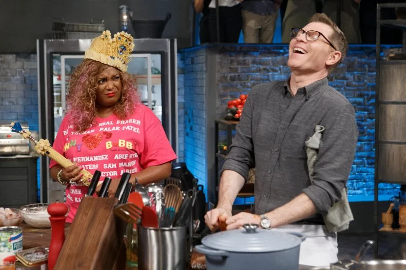 Co-host Sunny Anderson wears contender Francesco Buitoni's pasta crown and scepter as she checks in on chef Bobby Flay while he cooks Seafood Fra Diavolo during round two, as seen on Beat Bobby Flay, Season 21.