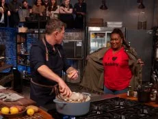 Co-host Sunny Anderson shows Bobby Flay an “I Love Bobby” t-shirt beneath her jacket as he cooks during round two, as seen on Beat Bobby Flay, Season 30.